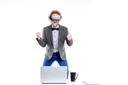 Portrait of curly-haired elegant businessman in vr glasses with hand in fist on chair at table with...