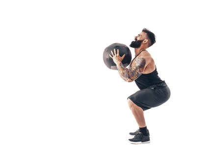Muscular tattooed bearded male exercising fitness weights Medicine Ball in studio isolated on white...