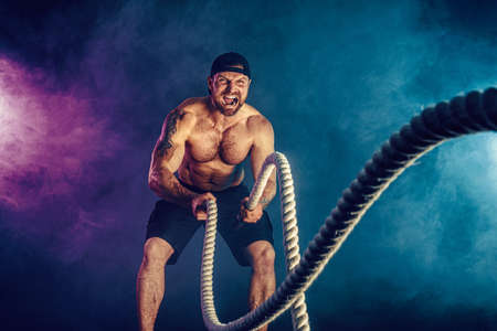 Bearded athletic looking bodybulder work out with battle rope on dark studio background with smoke. Strength and motivation.の写真素材