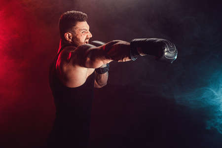 Bearded tattooed sportsman muay thai boxer in black undershirt and boxing gloves fighting on dark...
