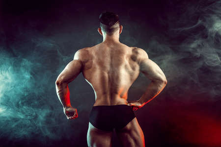Athletic man flexing muscles in studio on dark background with smoke. Strong bodybuilder with perfect abs.の写真素材