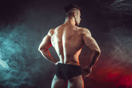 Athletic man flexing muscles in studio on dark background with smoke. Strong bodybuilder with perfect abs.の写真素材