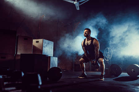 Muscular attractive caucasian bearded tasttoed man lifting kettlebell in a gym. Weight plates, dumbbell and tires in smoke background.の写真素材
