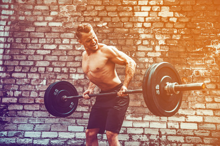 Athletic man working out with a barbell in front of brick wall. Strength and motivation. Outdoor workout. Exercise for the muscles of the backの写真素材