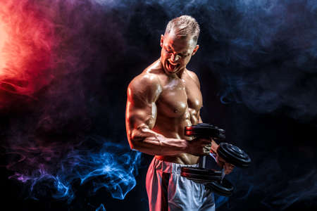 Topless man exercising biceps with dumbbells posing in studio full of colored smokeの写真素材