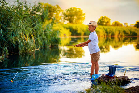 Little boy in straw hat fishing in lake at sunset.