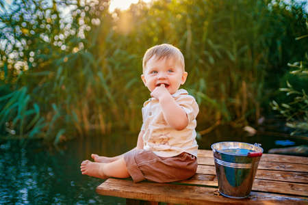 Little funny boy sitting on the edge of a wooden dock in lake at sunset.