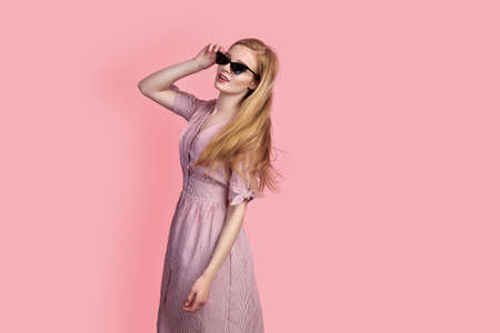Beautiful girl wearing dress and sunglasses posing, spinning around itself on pink background in studioの写真素材