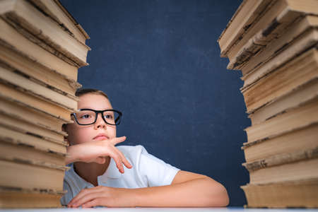 Smart boy in glasses sitting between two piles of books and look away thoughtfully.