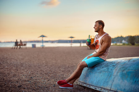 Young handsome sporty man in bright clothes sits on the seashore, drink a refreshing cocktail and enjoys his vacation.の写真素材