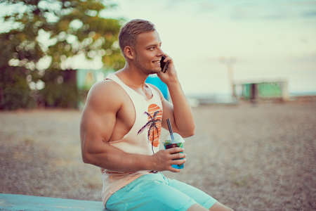 Young handsome sporty man in bright summer clothes sits on the seashore, drink a refreshing cocktail, speaking, chating on mobile phone and enjoys his vacation.の写真素材