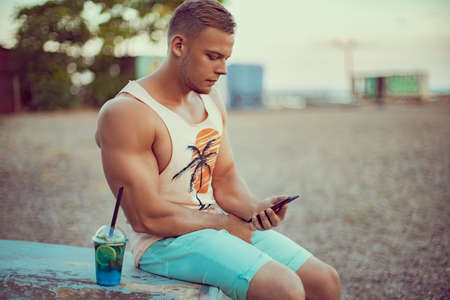 Young handsome sporty man in bright summer clothes sits on the seashore, drink a refreshing cocktail, speaking, chating on mobile phone and enjoys his vacation.の写真素材