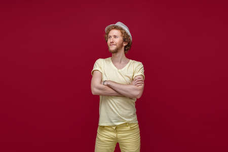 Fashionable hipster man posing with his arms crossed in studio. Smiling man in straw hat looking away from camera isolated in red backgroundの写真素材