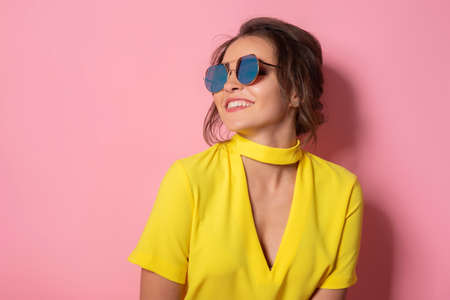 Beautiful girl in yellow dress wearing sunglasses posing, smiling on pink background in studio.の写真素材