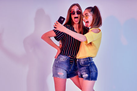 Two teenage girls in summer clothes standing in studio and making selfie on white background. Mixed...