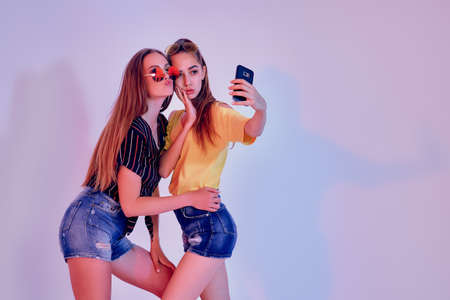 Two teenage girls in summer clothes standing in studio and making selfie on white background. Mixed...