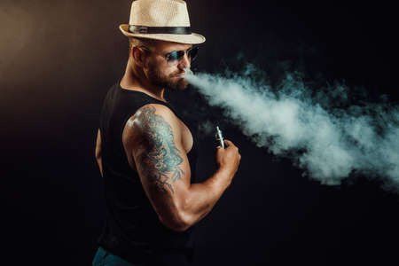 Bearded brutal male in hat and sunglasses smoking a vapor cigarette as an alternative to tobacco....
