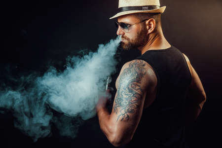 Bearded brutal male in hat and sunglasses smoking a vapor cigarette as an alternative to tobacco. Studio shot on dark background.の写真素材