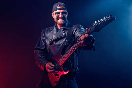 Brutal bearded Heavy metal musician in leather jacket and sunglasses is playing electrical guitar. Shot in a studio on dark background with smokeの写真素材