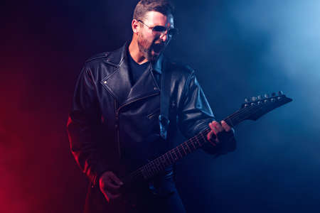 Brutal bearded Heavy metal musician in leather jacket and sunglasses is playing electrical guitar. Shot in a studio on dark background with smokeの写真素材