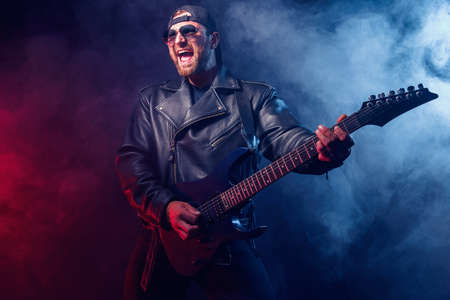 Brutal bearded Heavy metal musician in leather jacket and sunglasses is playing electrical guitar. Shot in a studio on dark background with smokeの写真素材