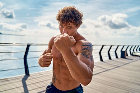 Muscular young man athlete standing and practicing shadow boxing outdoors early in the morning on pier by the seaの写真素材