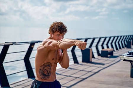 Muscular young man athlete standing and practicing shadow boxing outdoors early in the morning on pier by the seaの写真素材