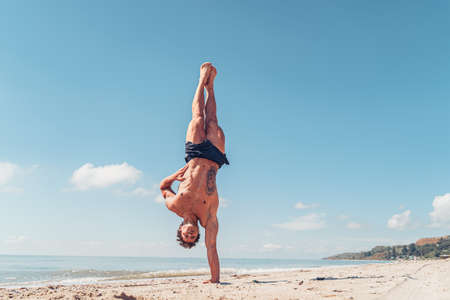 Muscular bodybuilder fitness man with a naked torso stands on one arm on the beachの写真素材