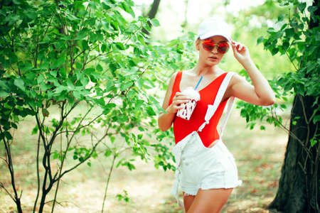 Portrait of sexy stylish fashion girl wearing rounded sunglasses, red swimsuit, white baseball cap, shorts with milkshake outdoors.の写真素材