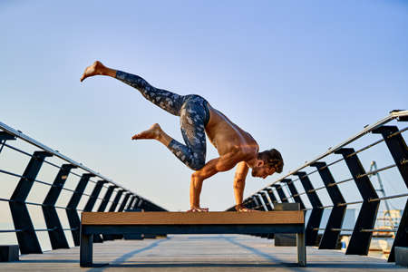 Fit man doing a hand stand while practicing yoga alone near the ocean against sky at dusk or dawn