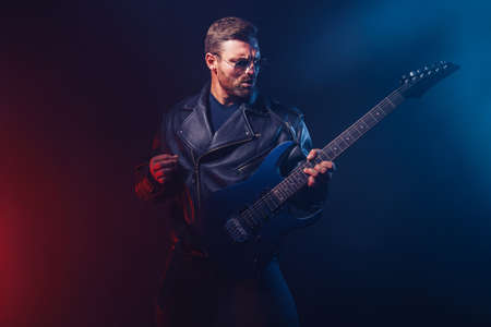Brutal bearded Heavy metal musician in leather jacket and sunglasses is playing electrical guitar. Shot in a studio on dark background with smokeの写真素材