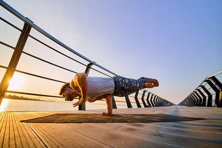 Fit man doing a one hand stand while practicing yoga alone near the ocean against sky at dusk or dawnの写真素材