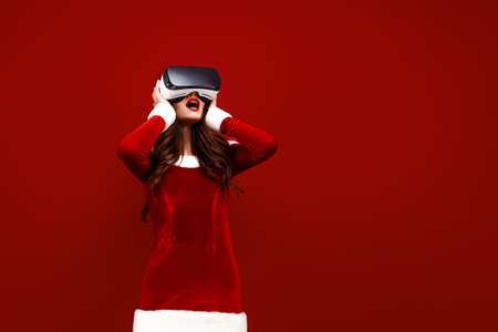 Amazed young Santa girl in Christmas dress looking in headset pointing hands aside isolated on red...