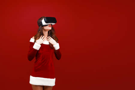 Amazed young Santa girl in Christmas dress looking in headset pointing hands aside isolated on red...