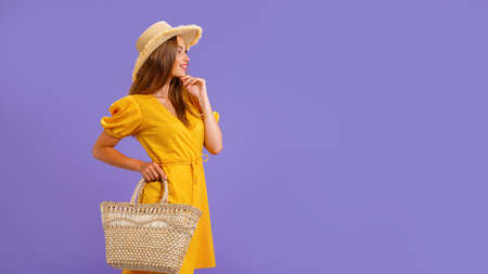 Beautiful smiling young woman in yellow dress, summer straw hat, bag isolated on pastel violet...