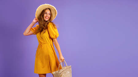 Beautiful smiling young woman in yellow dress, summer straw hat, bag isolated on pastel violet...