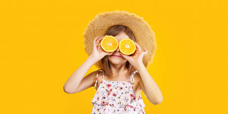 Happy little girl in a straw hat holding an orange on a yellow background with space for text.の写真素材