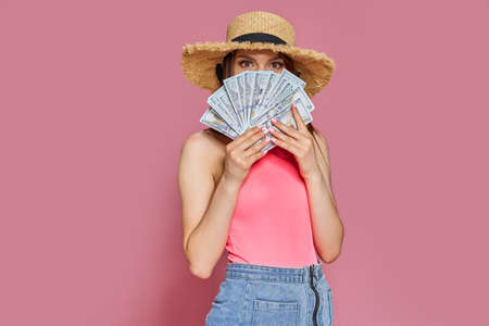 Smiling young tourist girl in straw hat hold fan of cash money in dollar banknotes isolated on pink...