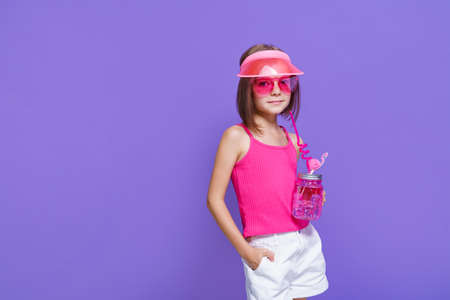a little girl in white shorts, a pink T-shirt, pink trendy glasses, with a drink in hand and a summer visor on a purple background in the studioの写真素材