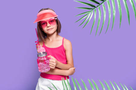 a little girl in white shorts, a pink T-shirt, pink trendy glasses, with a drink in hand and a summer visor on a purple background in the studioの写真素材