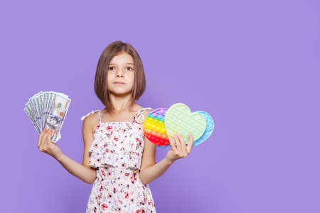 A little girl in a summer dress holds pop it in one hand, US dollar cash in the other and looking at...