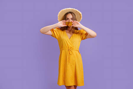 Cheerful young Caucasian woman in yellow dress and straw hat holding bank card isolated on purple...