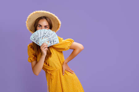 Charming lady in straw hat and yellow summer dress hold cash fan cover half face isolated purple...