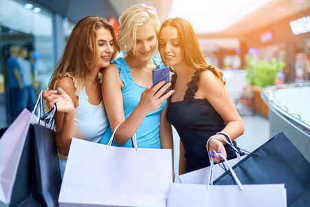 Three stylish woman holding shopping bags and look in mobile phone after shoppingの写真素材
