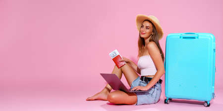Smiling girl with laptop tickets money and passport are going to travel. Sitting near suitcase in shorts white top and straw hat. Pink background. looks awayの写真素材