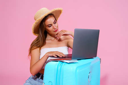 girl with laptop tickets money and passport are going to travel. Sitting near suitcase in shorts white top and straw hat. Pink background. looking at laptop. Something searchの写真素材