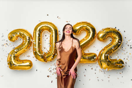 Happy young brunette woman near golden 2022 balloons on white background. New Year celebration....
