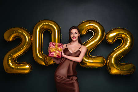 Georgeous brunette lady in beautiful dress holding gift box on black studio background with golden 2022 balloons, panorama. Happy young woman with wrapped present at New Year partyの写真素材