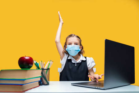Schoolgirl raising hand up and sitting at laptop wearing mask. Yellow background. Distance Learning