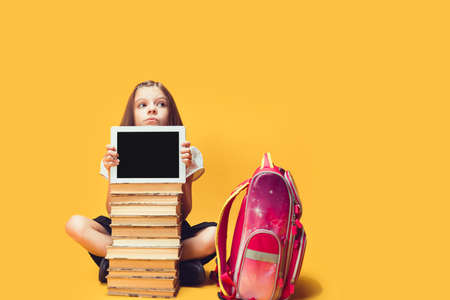 Thoughtful schoolgirl looks out from behind a stack of books and tablet. Kids education concept